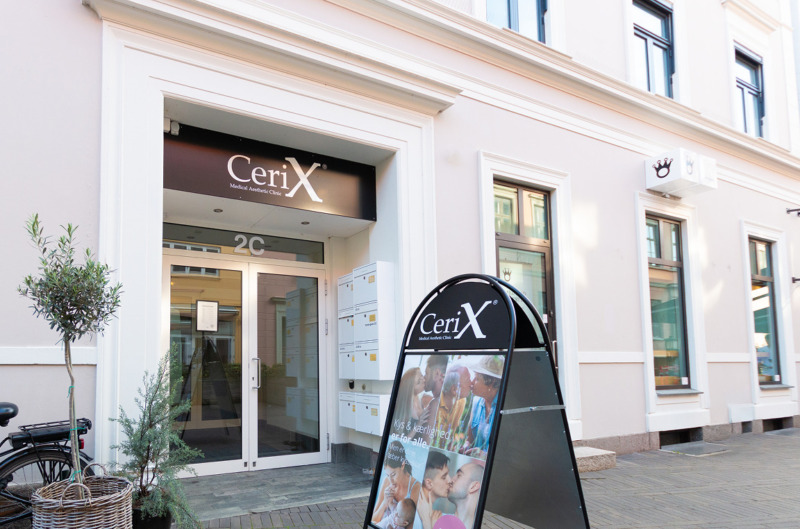 Cerix klinikfacade med indgangsdør, logo over døren og et udendørs reklameskilt. Skiltet viser billeder af ansigter og par – reklame for ansigtsbehandlinger som Botox. Billedet viser bygningens ydre, indgang og markedsføring af Cerix som æstetisk klinik. Gadescenen inkluderer vinduer, postkasser og planter ved indgangen.