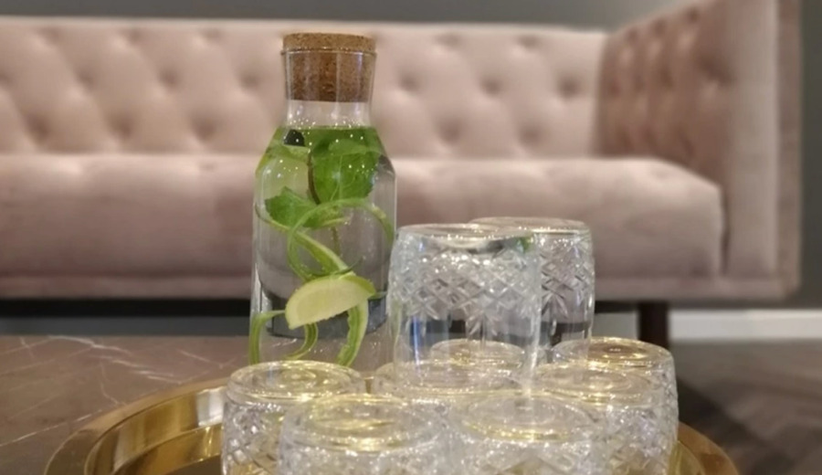 Billede fra Cerix’ venteværelse viser en klar glaskaraffel med skåret lime, mynte og korkprop samt flere vendte krystalglas på en guldbakke foran en rosa chesterfield-sofa. Stemningsfuldt, serviceorienteret foto relateret til behandlinger hos Cerix, fx Botox til ansigtet, til besøgende og patienter.