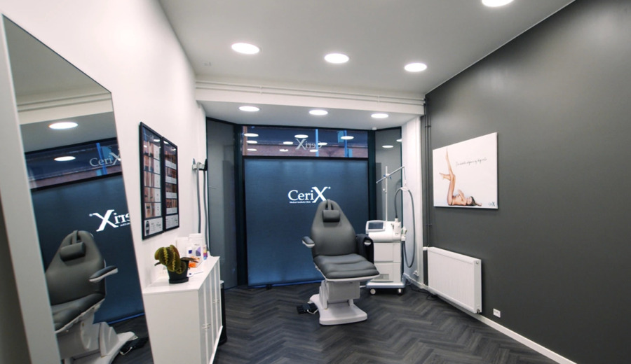 Behandlingsrum hos Cerix med en justerbar komfortabel patientstol, lasersystem, instrumentvogn og dekorativt spejl. Moderne, sterilt udseende med mørke vægge, loftslamper, hvidt skab, fiskeben-gulv og vinduesfolie med Cerix-logo. Billedet illustrerer en Botox-behandling rettet mod ansigtet (ansigt), klar til behandling uden patient.