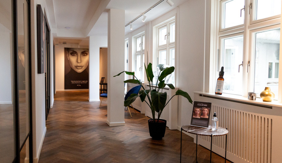 Lyse, moderne venteområde hos Cerix med trægulv, store vinduer, grøn plante, siddepladser og plakater. Plakat viser ansigt og reklamerer for Botox-behandling af ansigtet. Bord med produktflaske og før/efter-brochure signalerer klinikkens fokus på ansigtsbehandlinger. Lysindfaldet skaber en rolig atmosfære.