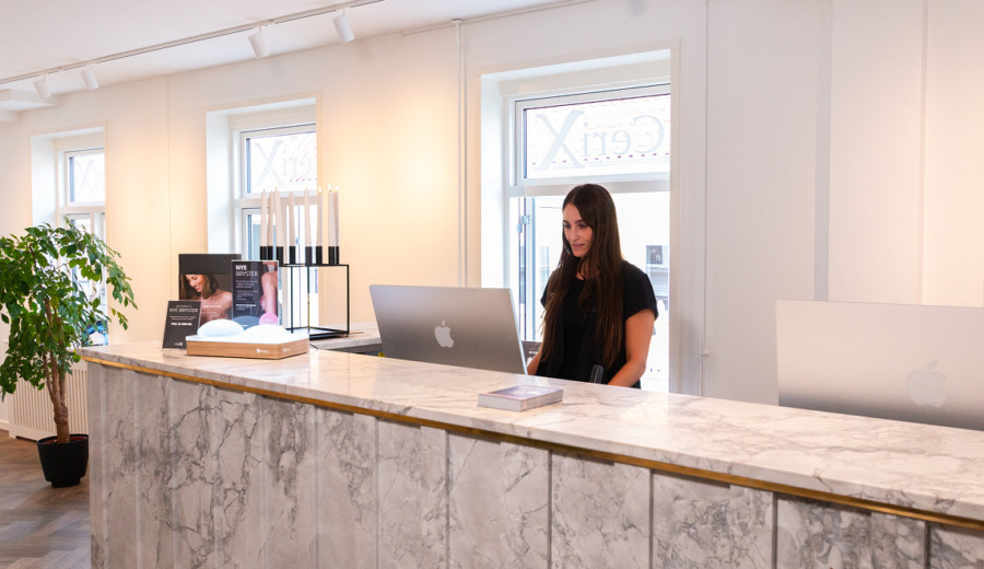 Receptionen hos Cerix med en kvindelig receptionist bag en marmorbetonet skranke, computerskærm og informationsmateriale. Brochurer viser tilbud om brystforstørrende operation (BFO) med billeder af bryster, samtidig ses receptionistens ansigt. Personalet modtager klienter i et roligt, lyst og stilrent, veludstyret miljø.