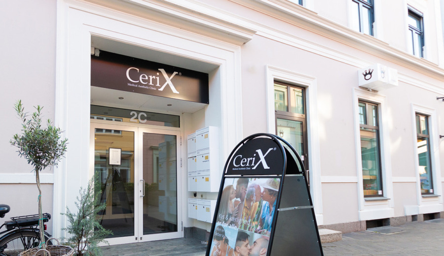 Billede af Cerix Medical Aesthetic Clinics lyse facade og glasindgang i en bygade, med et fritstående reklameskilt foran. Skiltet viser nærbilleder af ansigter; Cerix fremhæver behandlinger som Botox og ansigtsbehandlinger ved klinikkens indgang. Nummeret 2C over døren, postkasser, cykel og planter på fortovet.