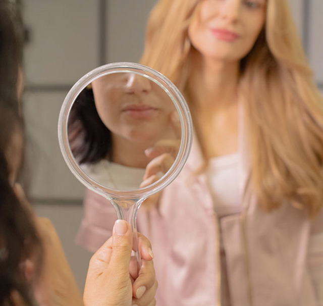Cerix: En hånd holder et rundt spejl, der reflekterer en anden kvindes underansigt med læber og hage, mens en blond kvinde ses sløret i baggrunden. Billedet illustrerer en klinisk konsultationssituation med fokus på ansigtsbehandlinger hos Cerix, for eksempel Botox eller filler.