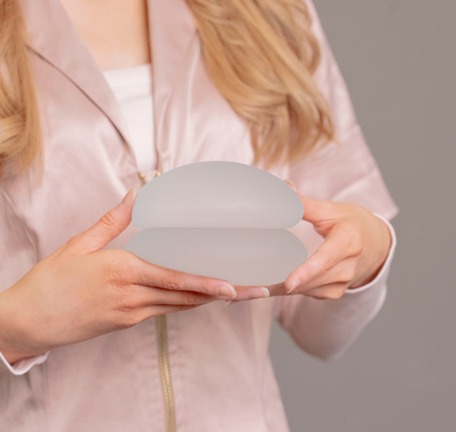 Person holder to silikonebrystimplantater foran kroppen i et tæt crop; hænderne og implantaterne er i fokus, ryggen er dækket af en lyserød bluse. Billedet bruges af Cerix som illustration til brystforstørrende operation (BFO) og viser brystrelateret behandling med neutral grå baggrund og tydelige fingerspidser.