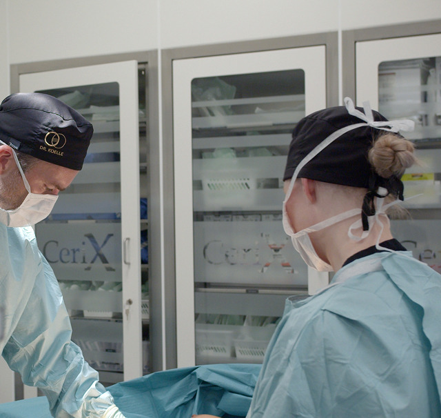 Operatørteam i Cerix‑kirurgisk lokale iført operationskitler, kasketter og masker under en brystforstørrende operation (BFO). To læger og en sygeplejerskeassistent står ved patientens dækkede brystområde, arbejder med sterilt blåt dække og instrumenter; Cerix-logo ses på skabe i baggrunden, fokuseret under operationslamper i sterile forhold.