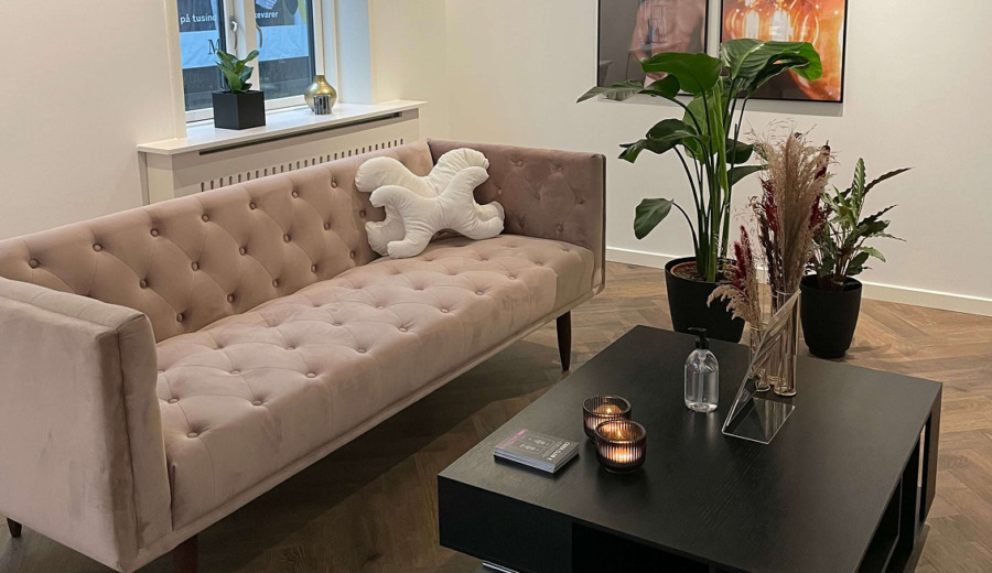 Moderne, roligt venteområde hos Cerix i nordisk stil med rosa chesterfield-sofa, hvid dekorativ pude, vindueskarm med pynt, sort sofabord med tændte stearinlys, håndsprit, informationsbrosjyre og magasiner, grønne planter, plakater på væggen og varmt trægulv. Klinikken tilbyder blandt andet professionelle Botox-behandlinger til ansigtet.