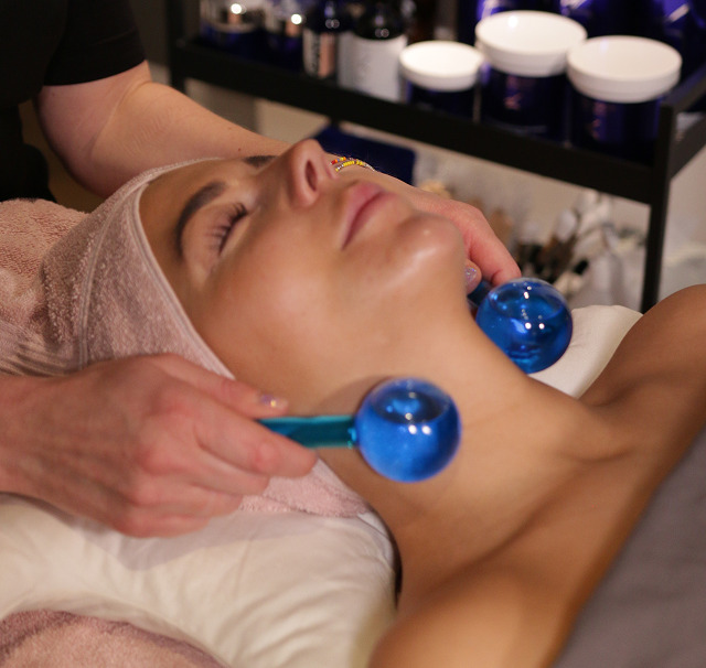 Cerix-klinik: En kvinde ligger med lukkede øjne og håndklæde om håret under en ansigtsbehandling. En behandler bruger to blå massageredskaber til at arbejde på ansigt og hals. Billedet viser en professionel, afslappet ansigts- og halsmassage med hudplejeprodukter i baggrunden hos Cerix.