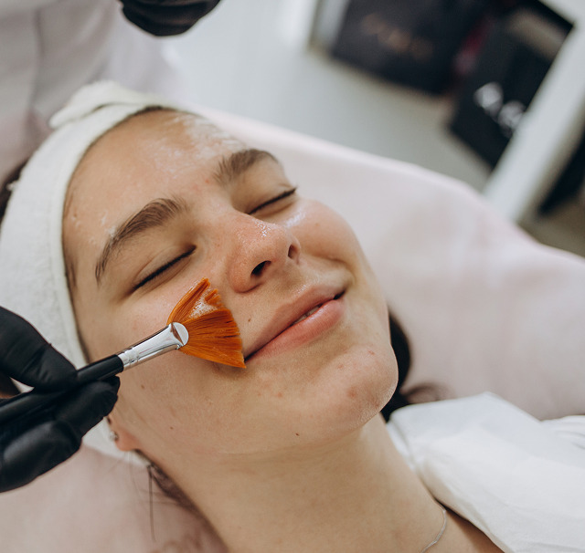 Cerix-klinik: Kvindelig patient ligger afslappet med lukkede øjne mens en behandler påfører en orange ansigtsmaske med blød pensel. Billedet viser en kosmetisk ansigtsbehandling (ansigtsmaske) fokuseret på ansigtet og hudpleje i en professionel, klinisk setting med fokus på rens, eksfoliering og beroligelse af huden.