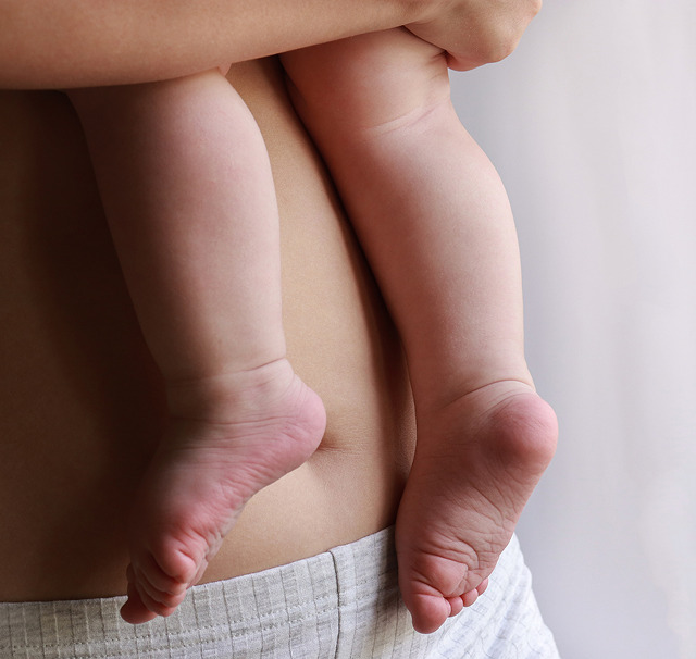 Cerix: Billede af en voksen, der holder et spædbarn placeret på hoften, med bare babys ben og fødder hvilende mod den voksnes mave og side. Billedet illustrerer maveområdet og kan bruges i forbindelse med Maveplastik eller kropskonturering hos Cerix, fx efter fødsel.