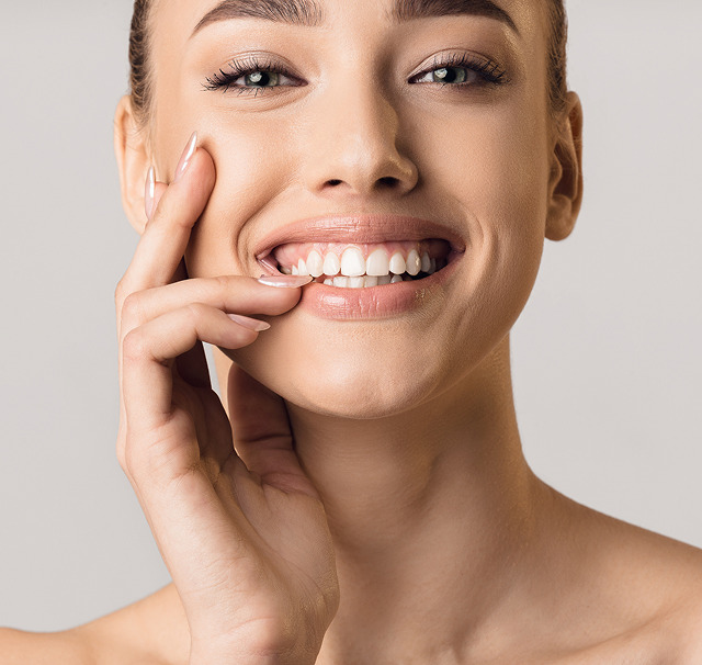 Nærbillede af en smilende kvinde der rører sin kind og viser hvide tænder; billedet fokuserer på glat hud, fyldige læber og ansigtets konturer, naturligt smil og klart blik. Cerix tilbyder ansigtsbehandlinger som Botox for at reducere rynker og fremhæve et friskt, ungdommeligt udseende.