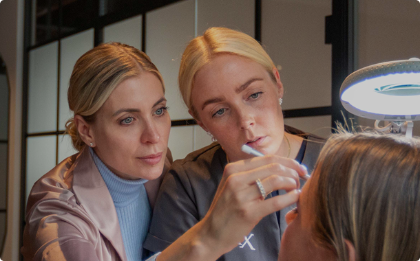 På billedet ses to behandlere i en Cerix klinik, der udfører en kosmetisk injektionsbehandling i en patients ansigt. Den ene behandler fokuserer nøje på området omkring patientens øje og bryn, understøttet af lys fra en luplampe, hvilket typisk indikerer en behandling med Botox eller fillers mod rynker.